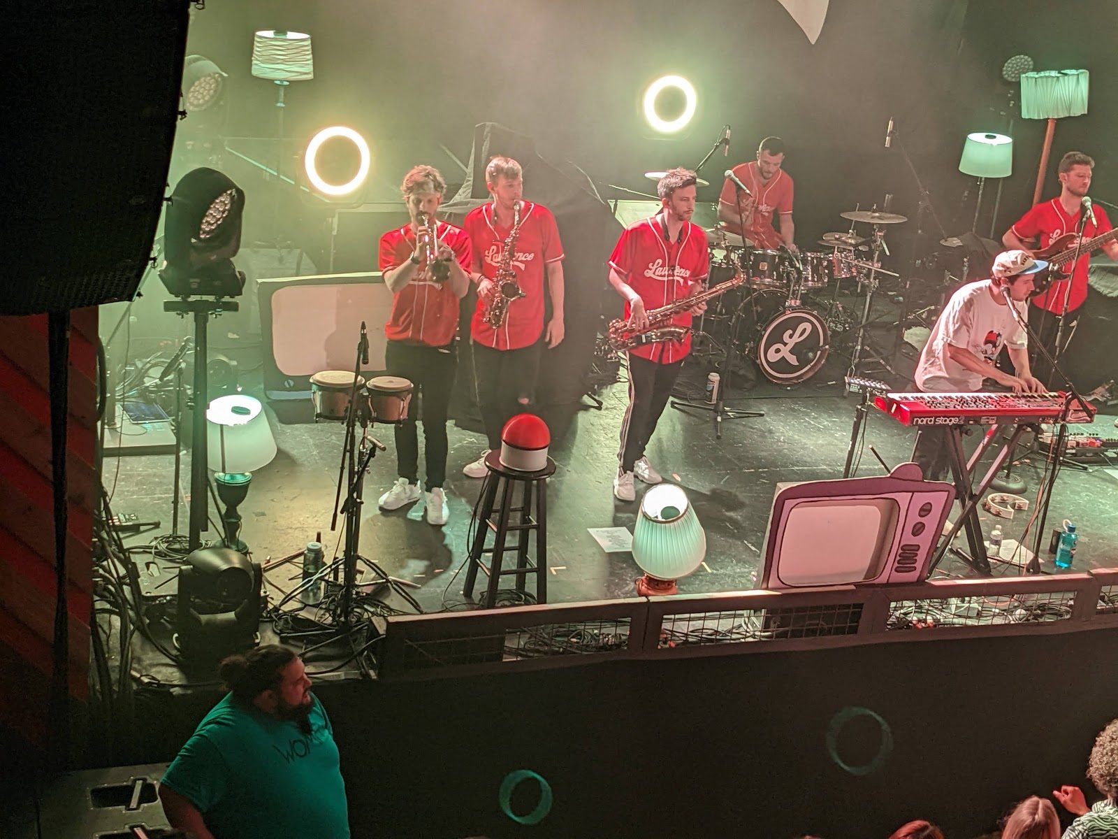 A band in red baseball jerseys playing trumpet, saxophones, drums and keyboards on an indoor White Oak Music Hall stage, seen from above the barrier rail