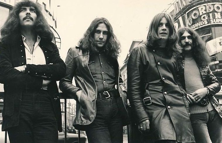 Black-and-white photo of the four members of Black Sabbath in long hair and coats, leaning on a railing on a city street