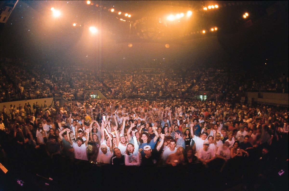 View from the stage of a packed arena floor, fans with raised arms under warm stage lights and full seating tiers behind
