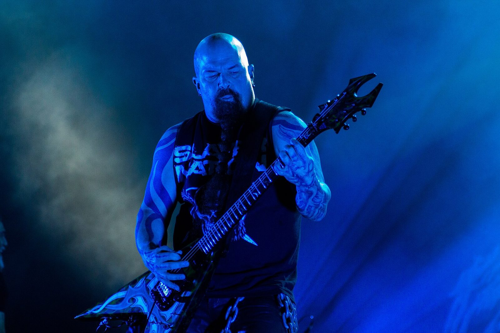 Kerry King, bald with a long beard and tattooed arms, playing a pointed V-shaped guitar under blue stage light and haze