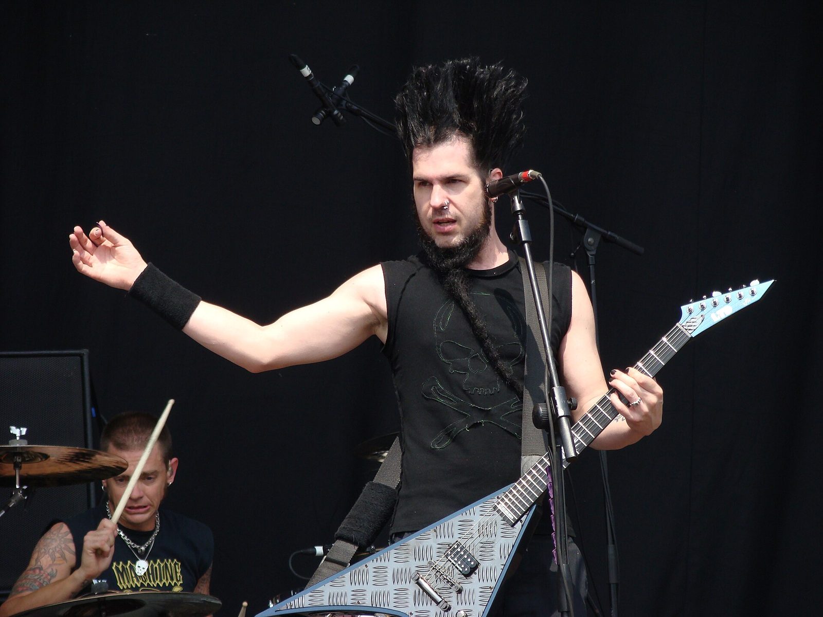 Wayne Static of Static-X, with tall spiked black hair and a long braided goatee, playing a diamond-plate V-shaped guitar at a microphone while a drummer plays behind him