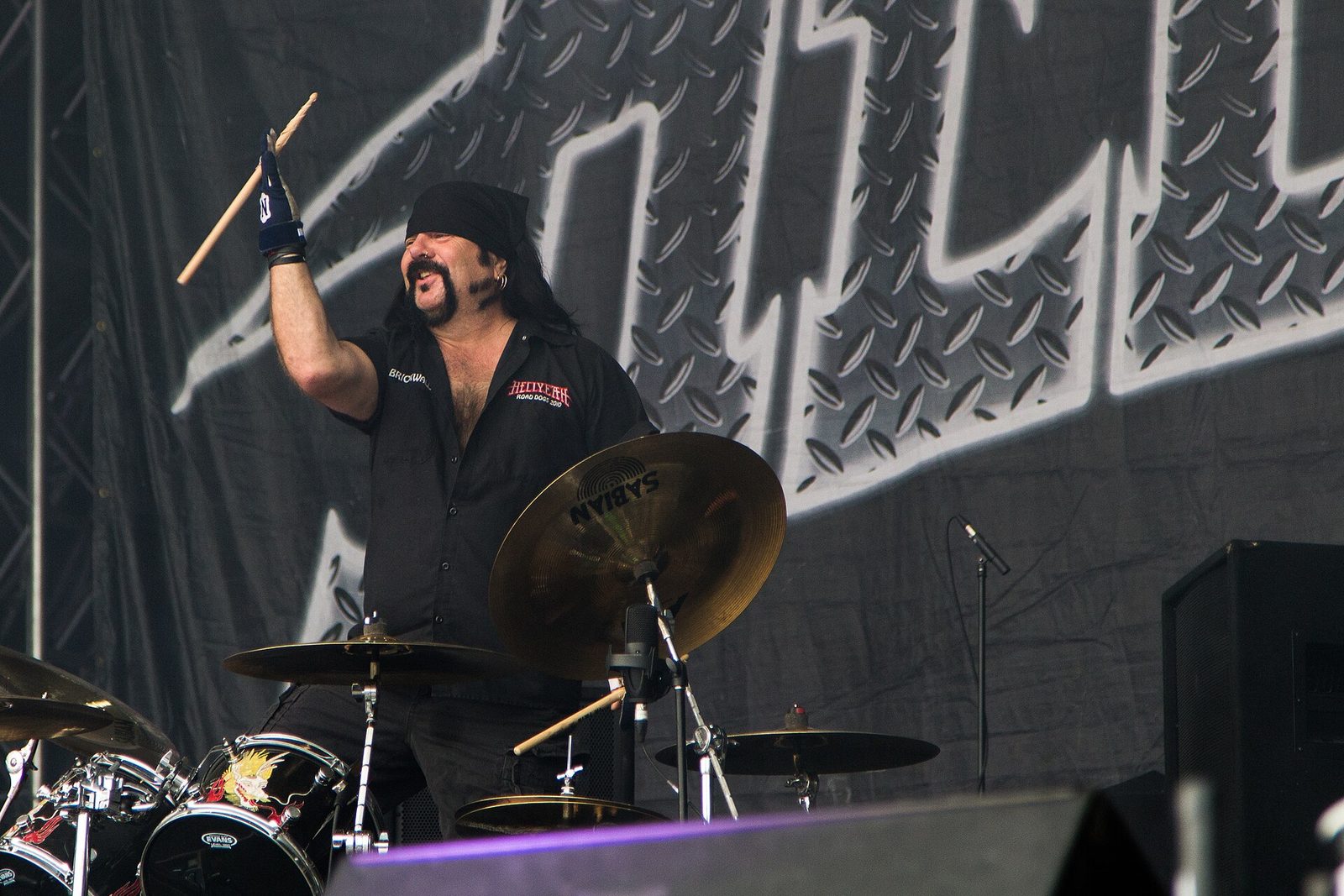 Vinnie Paul in a black bandana and black work shirt raising a drumstick behind his kit and a Sabian cymbal, in front of a large diamond-plate stage backdrop