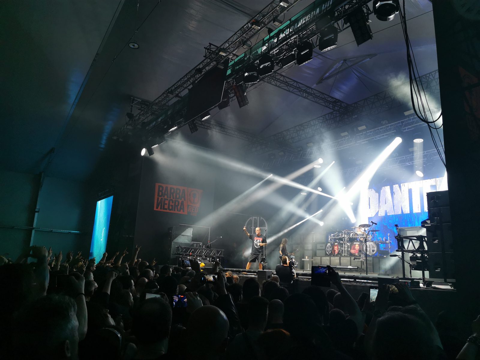 Pantera on a smoky stage in Budapest in 2023, Phil Anselmo raising a fist under white light beams, a long-haired player beside him and a crowd raising horns in the foreground