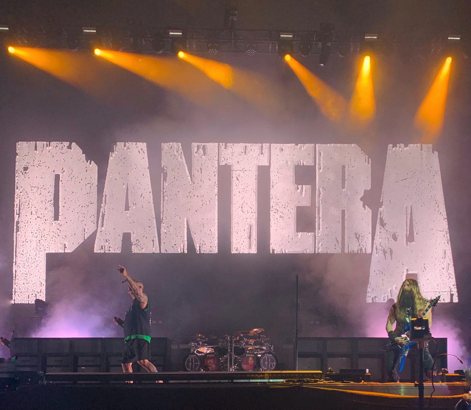 Pantera onstage at Copenhell in 2023 under orange spotlights, with a giant distressed white Pantera logo behind the drum kit, Phil Anselmo pointing at the crowd and a long-haired guitarist at right