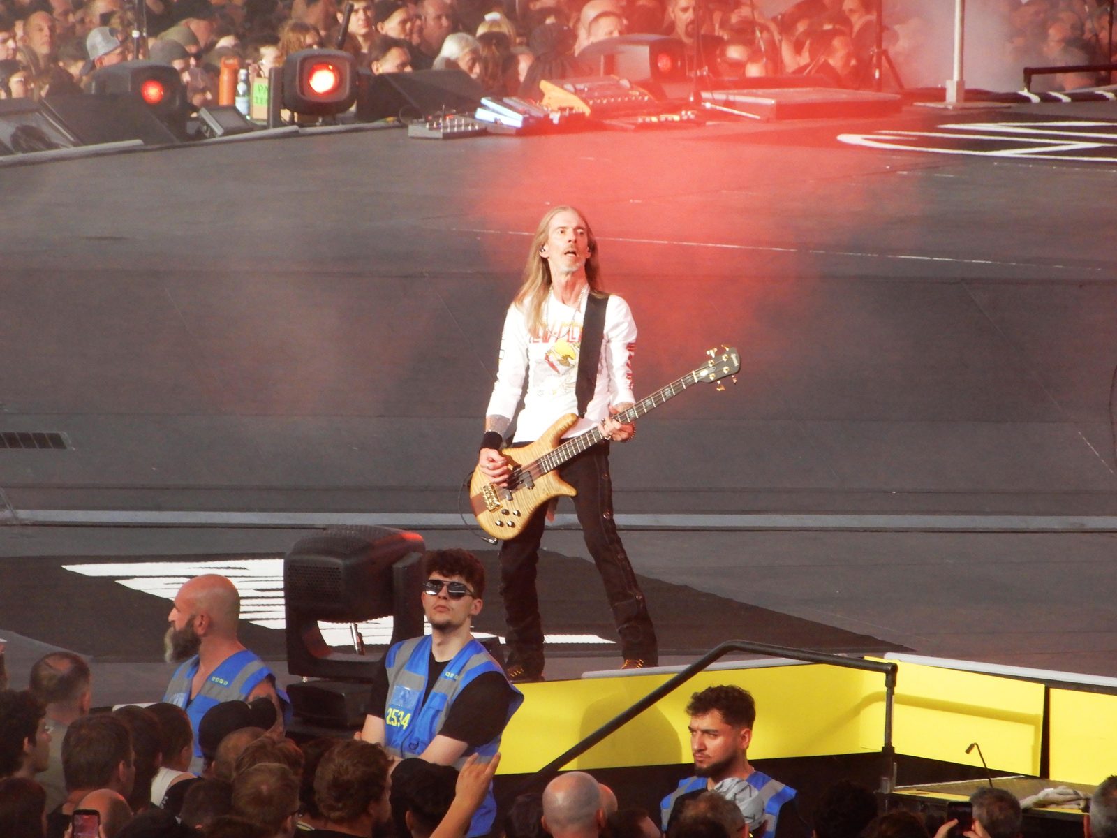 Rex Brown, with long grey-blond hair and a white long-sleeved shirt, playing a natural-wood bass on a stadium stage while security staff in blue vests stand in front of the crowd