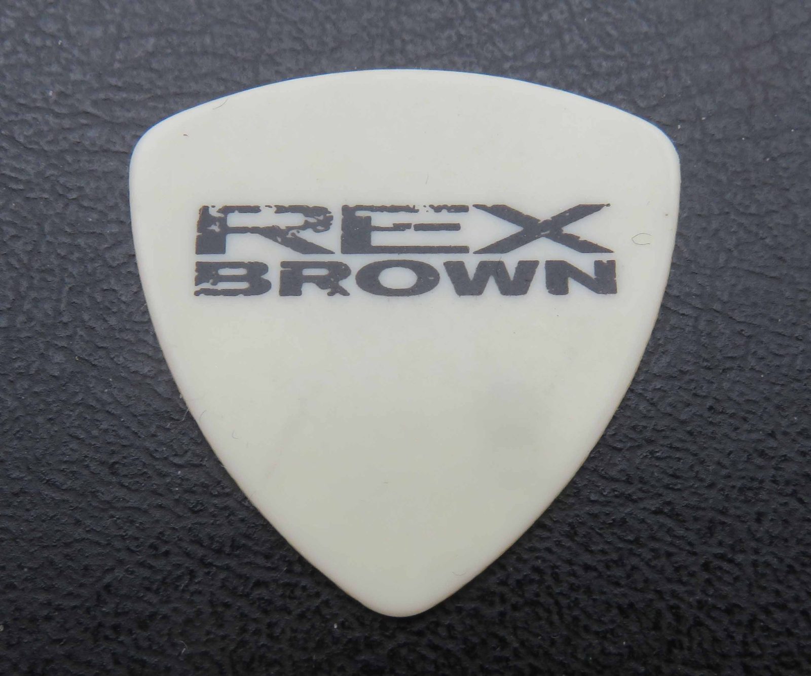 A cream-colored triangular guitar pick printed with REX BROWN in distressed black letters, lying on a black leather surface