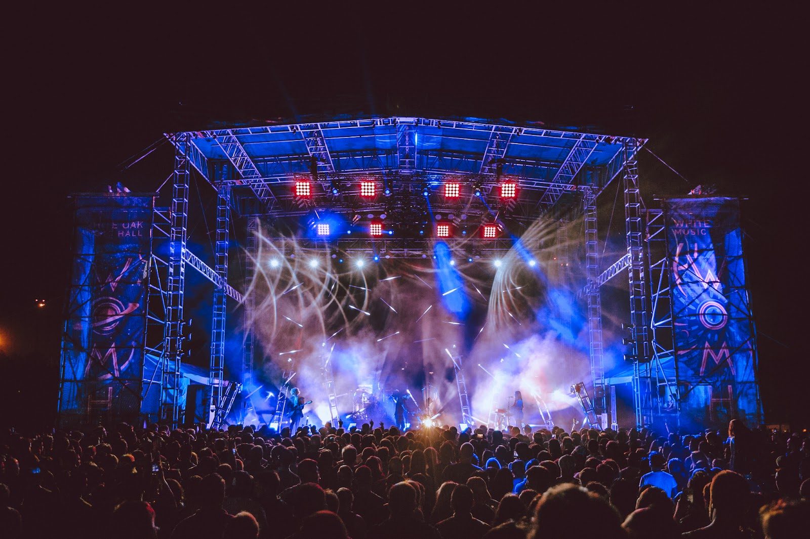 A night crowd packed in front of White Oak Music Hall's outdoor lawn stage, a steel truss roof lit blue and red with beams cutting through haze and venue banners on the side towers