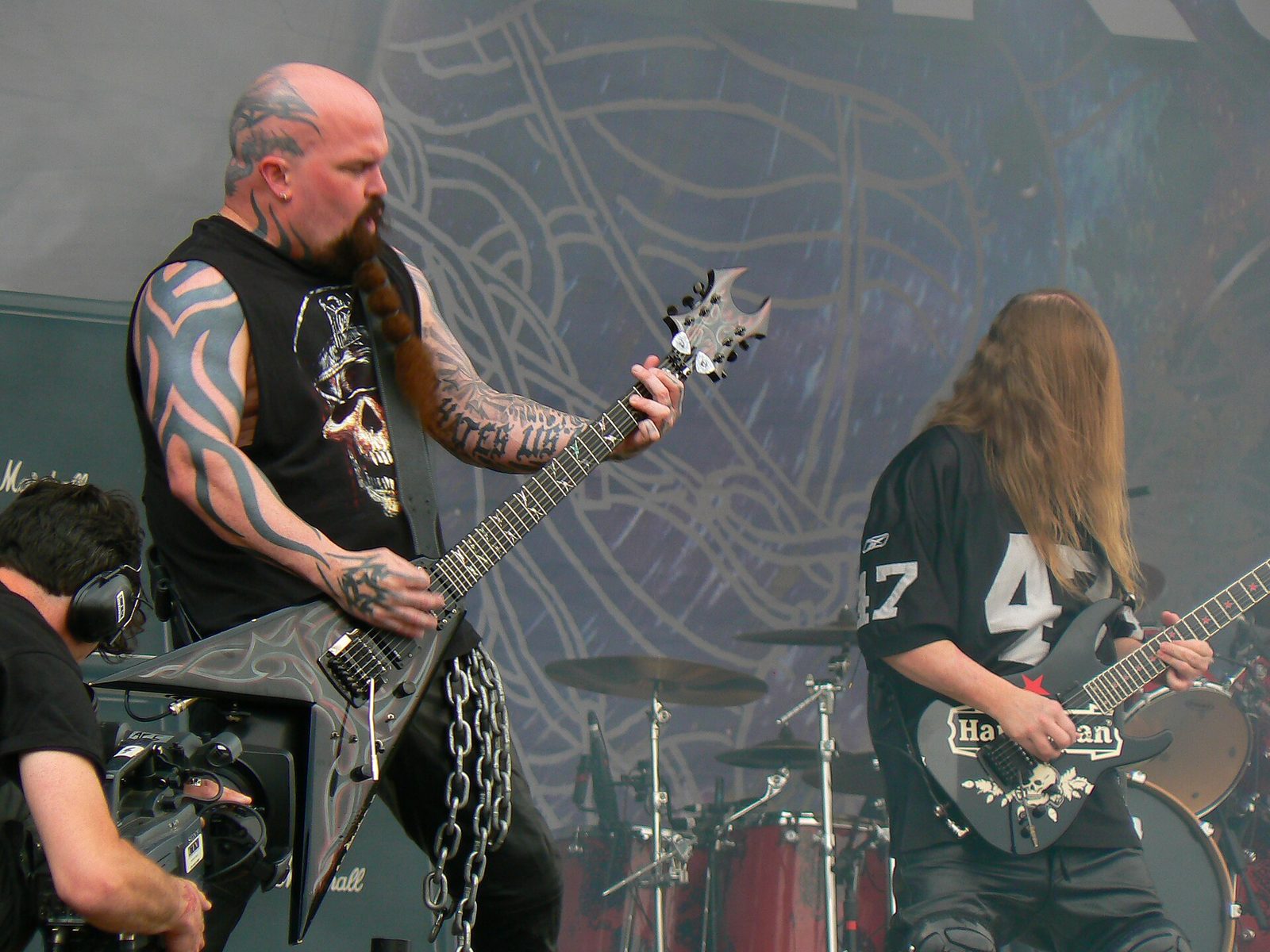 Kerry King, bald with tattoos on his head and arms and a long braided beard, playing a black V-shaped guitar beside Jeff Hanneman, whose long blond hair covers his face, in a black number 47 jersey
