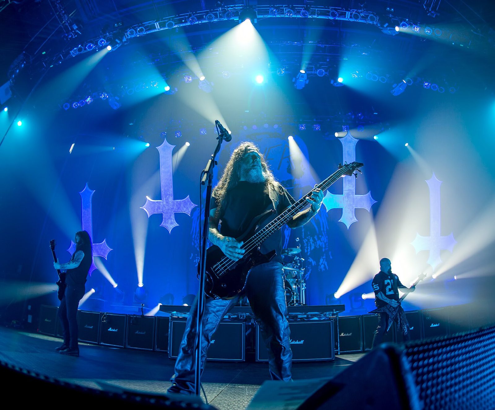 Tom Araya playing bass at a center microphone under blue and white stage lights, Gary Holt at left and Kerry King in a black football jersey at right, large inverted crosses lit on the backdrop