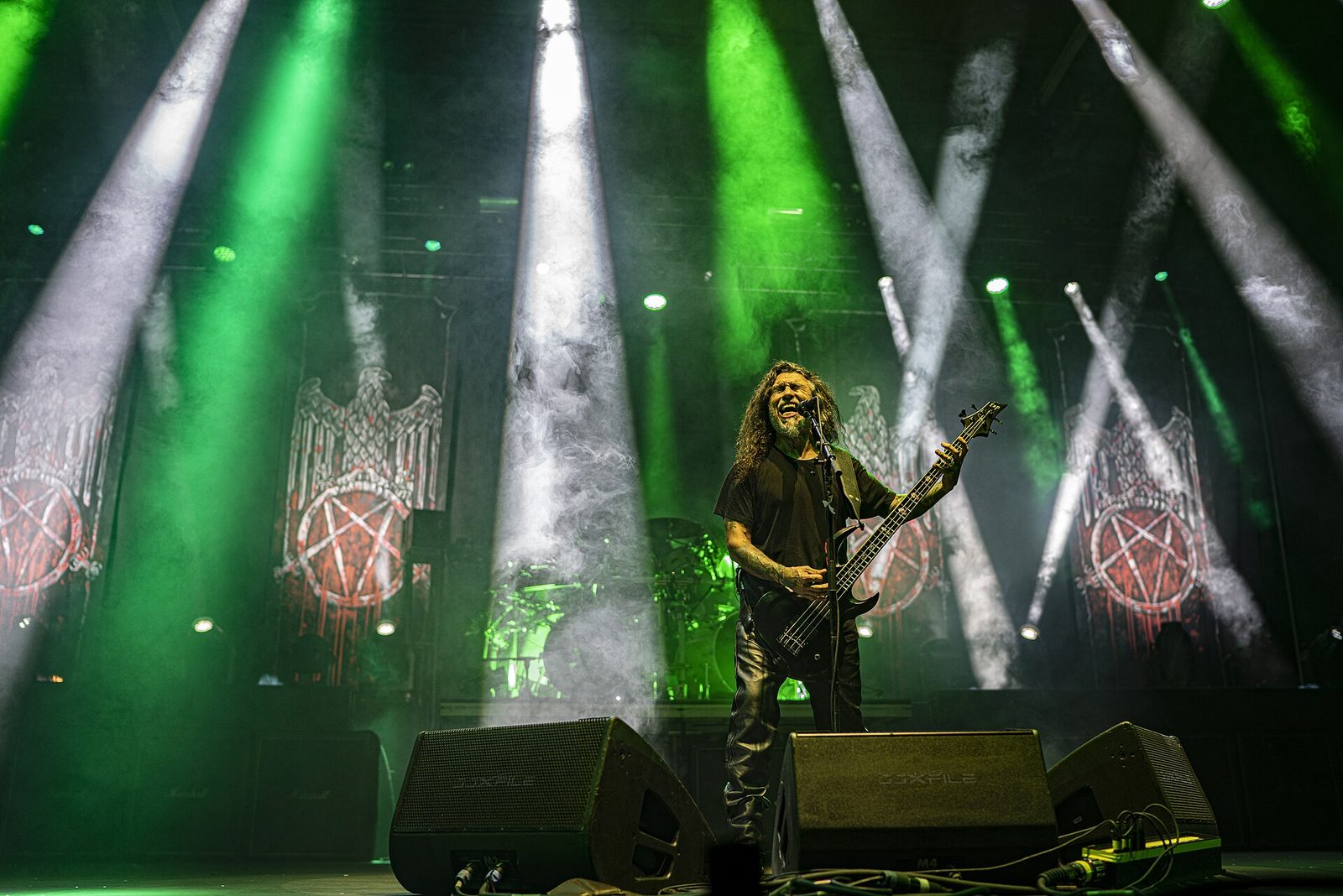 Tom Araya, long dark hair and a grey beard, singing into a microphone with his bass, alone at the front of a stage lit with green and white beams, eagle-and-pentagram banners behind him