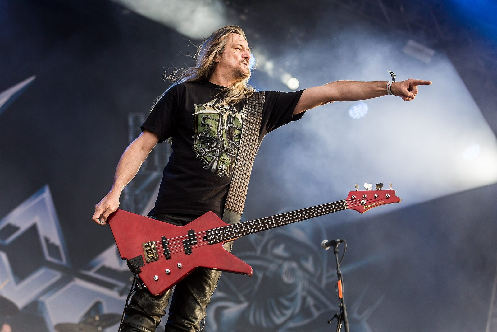 Tom Angelripper of Sodom on an outdoor festival stage, long blond hair, a black Sodom T-shirt and a red bass guitar, pointing out at the crowd with his left arm