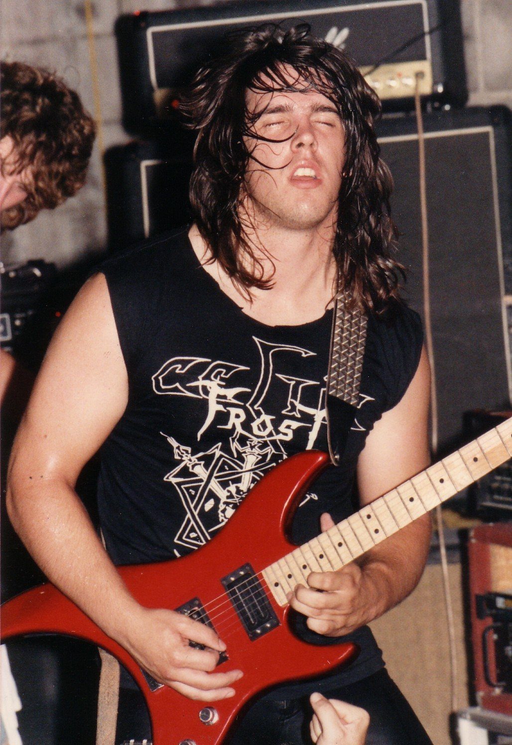 A young John Perez with long dark hair playing a red electric guitar, eyes closed, in a sleeveless Celtic Frost shirt in front of an amplifier stack