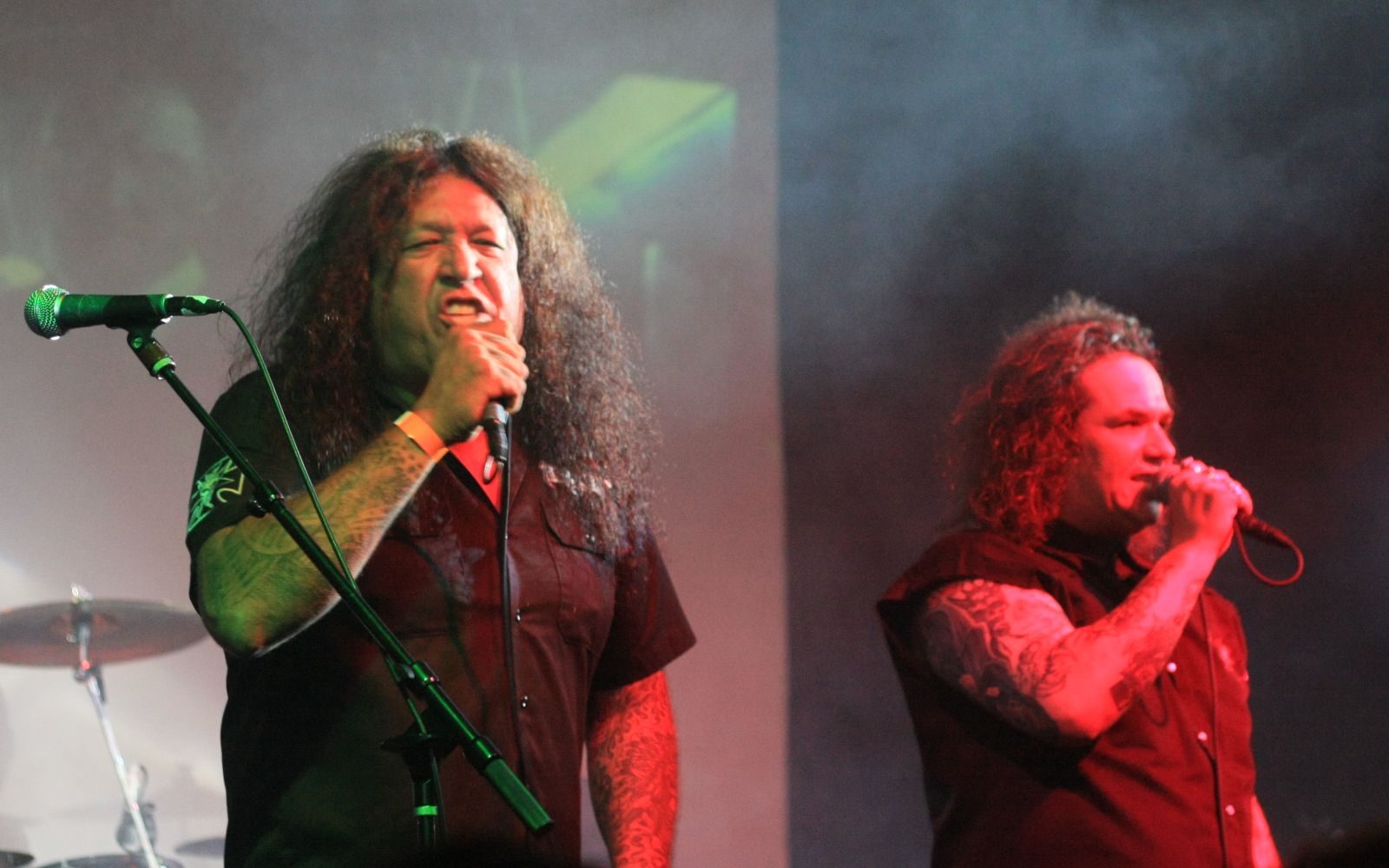 Chuck Billy, long-haired and tattooed, singing into a microphone at left while Steve Souza sings into a handheld microphone at right, both in dark shirts on a smoky stage
