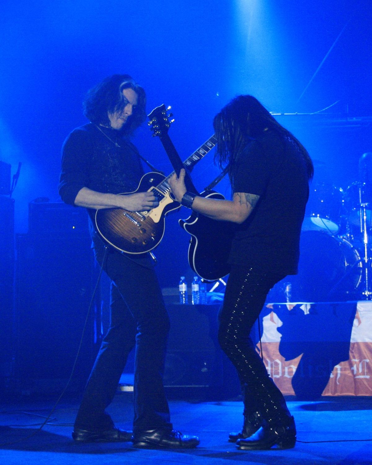 Alex Skolnick with a sunburst Les Paul and Eric Peterson with a black guitar playing face to face under blue stage light