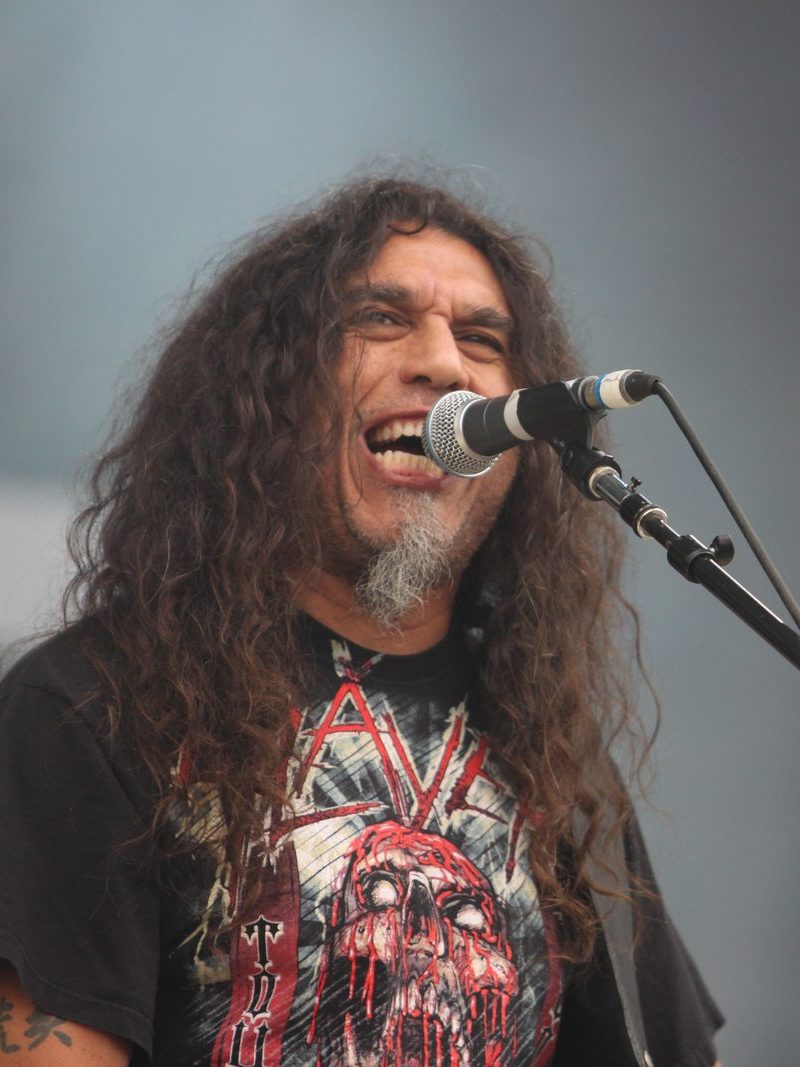 Slayer singer and bassist Tom Araya, with long curly hair and a gray goatee, singing into a microphone in a black Slayer T-shirt