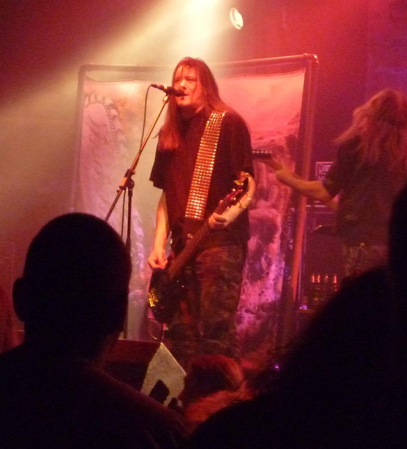 Tom Angelripper of Sodom singing into a microphone while playing bass, wearing a studded strap, under red and white stage light