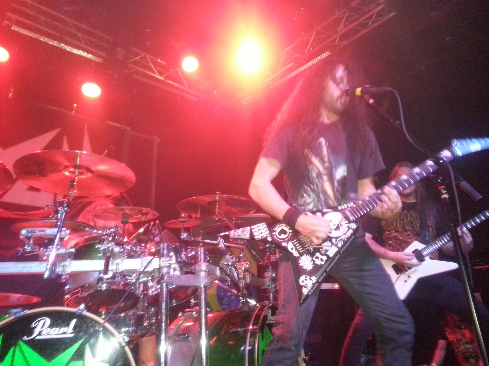 Havok onstage in 2013: a long-haired guitarist sings into a microphone while playing a sticker-covered black V guitar under red stage lights, a drum kit and second guitarist beside him