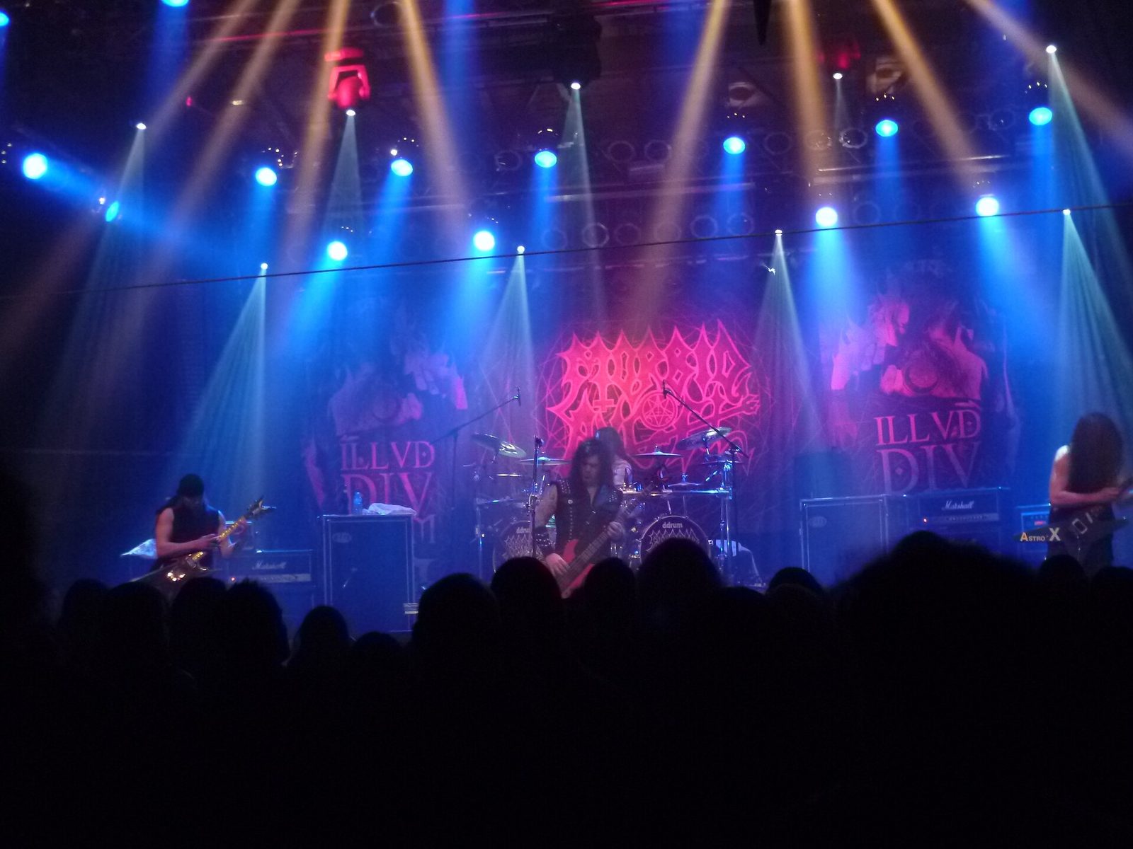 Morbid Angel on a club stage under blue and white spotlight beams, a bassist center stage and two guitarists at the edges, with the band's red logo on the backdrop and a dark crowd in the foreground