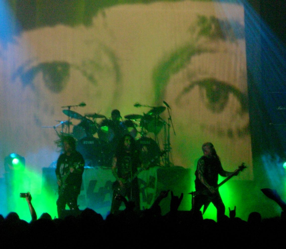 Slayer on a stage washed in green light, two guitarists and a bassist in silhouette in front of a drum kit, with a huge projected black-and-white image of a pair of eyes behind them and raised hands in the crowd