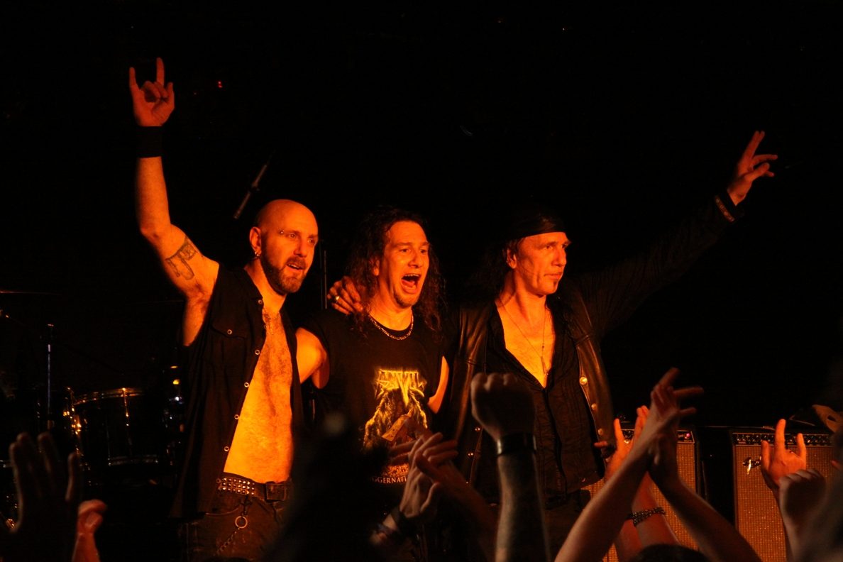 Three members of Anvil standing together at the front of a dark stage under orange light, two of them throwing horns, with fans' raised hands in the foreground