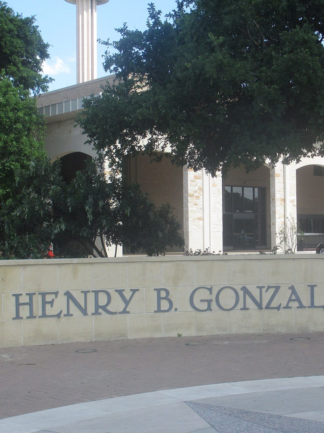 The arched stone front of the Henry B. Gonzalez Convention Center behind a live oak and a low wall lettered with its name, with the column of the Tower of the Americas rising above the roof