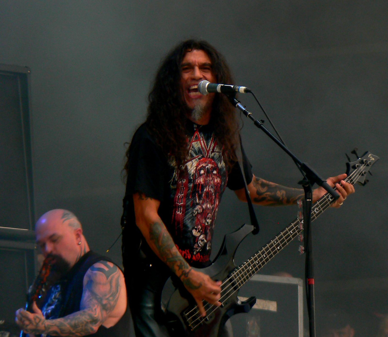 Tom Araya in a red-and-black Slayer T-shirt singing into a microphone and playing a black ESP bass, with Kerry King, bald with tattooed arms, playing guitar in the foreground at left