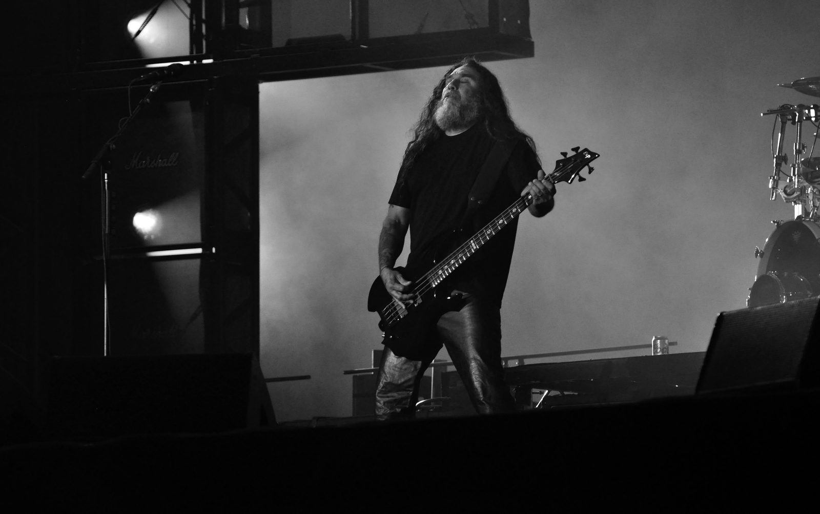 Black-and-white photo of Tom Araya, with long hair and a grey beard, head tilted back and eyes closed, playing a dark bass on a smoky festival stage beside a Marshall speaker cabinet