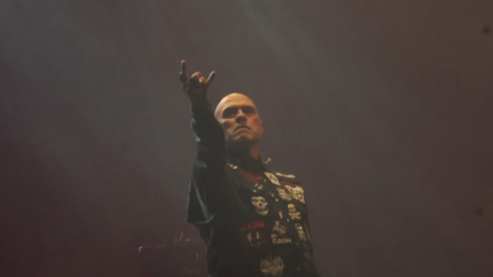 Vio-lence singer Sean Killian, bald and wearing a black vest covered in band patches, raising devil horns toward the stage lights through haze