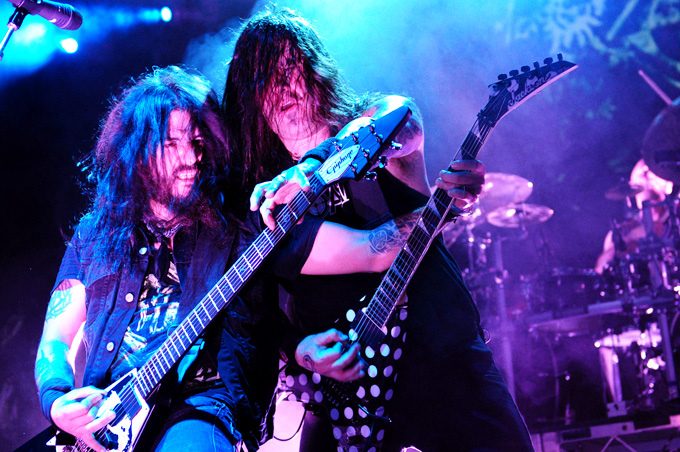 Robb Flynn and Phil Demmel standing shoulder to shoulder under blue stage lights, both with long dark hair, one playing a black Epiphone guitar and the other a black guitar with white polka dots